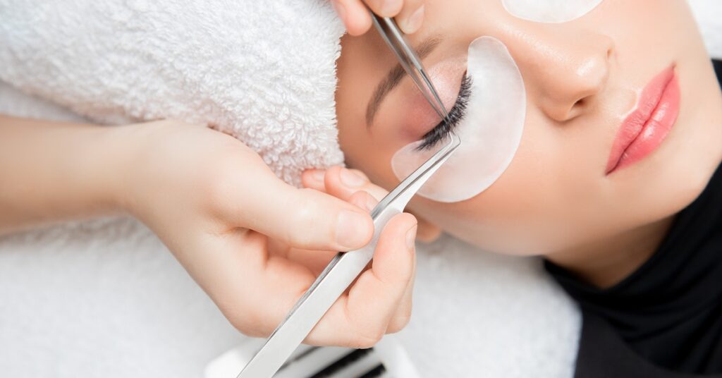 A pair of hands performing an eyelash extension procedure on a woman's eyelashes with a pair of tweezers.