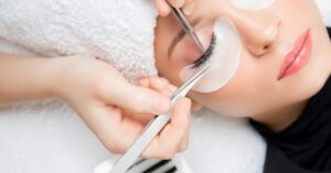 A pair of hands performing an eyelash extension procedure on a woman's eyelashes with a pair of tweezers.