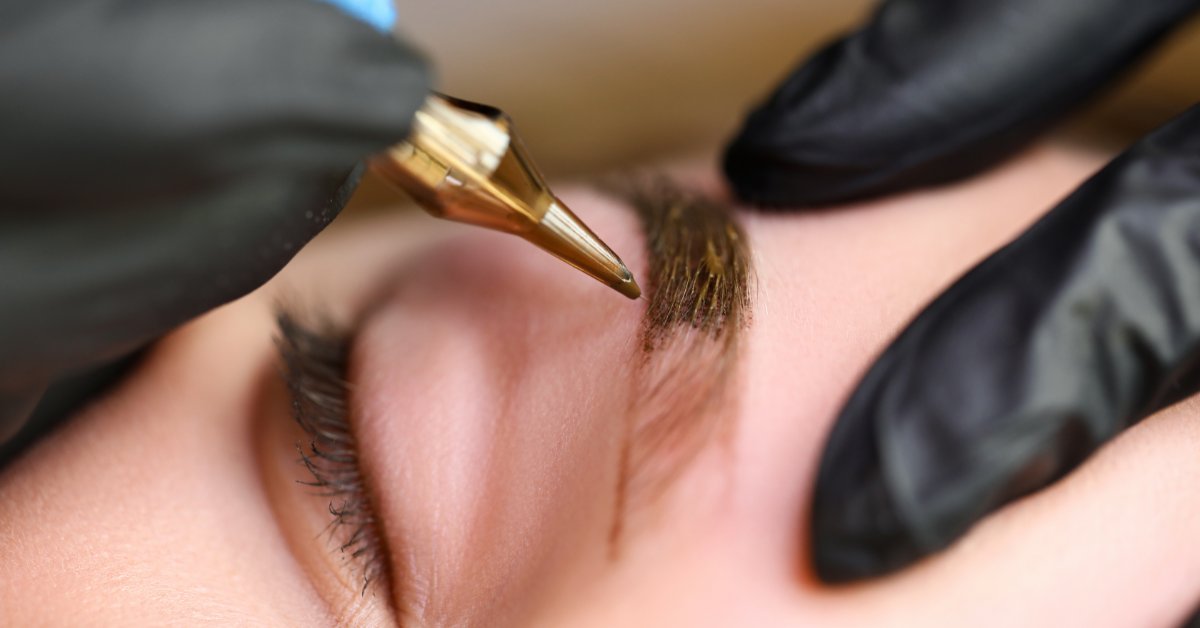 A closeup on a pair of gloved hands performing a permanent eyebrow tattoo procedure with a tattoo instrument.