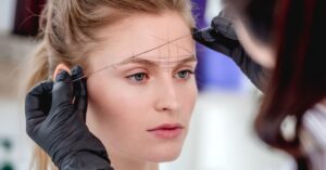 A cosmetologist wearing gloves is in the process of microblading a woman's eyebrows according to lines on her forehead.