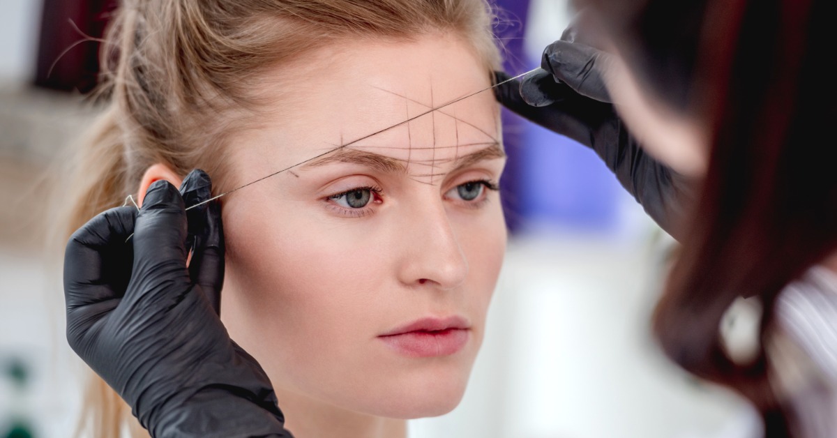 A cosmetologist wearing gloves is in the process of microblading a woman's eyebrows according to lines on her forehead.