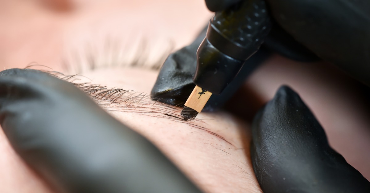 A closeup on a pair of gloved hands performing a microblading procedure on a person's eyebrows with a microblading tool.
