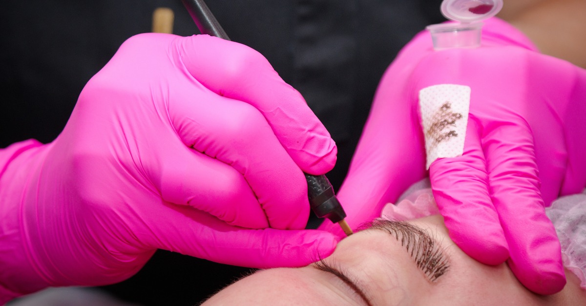 A makeup artist wearing pink rubber gloves performing a microblading procedure on a patient's eyebrows.
